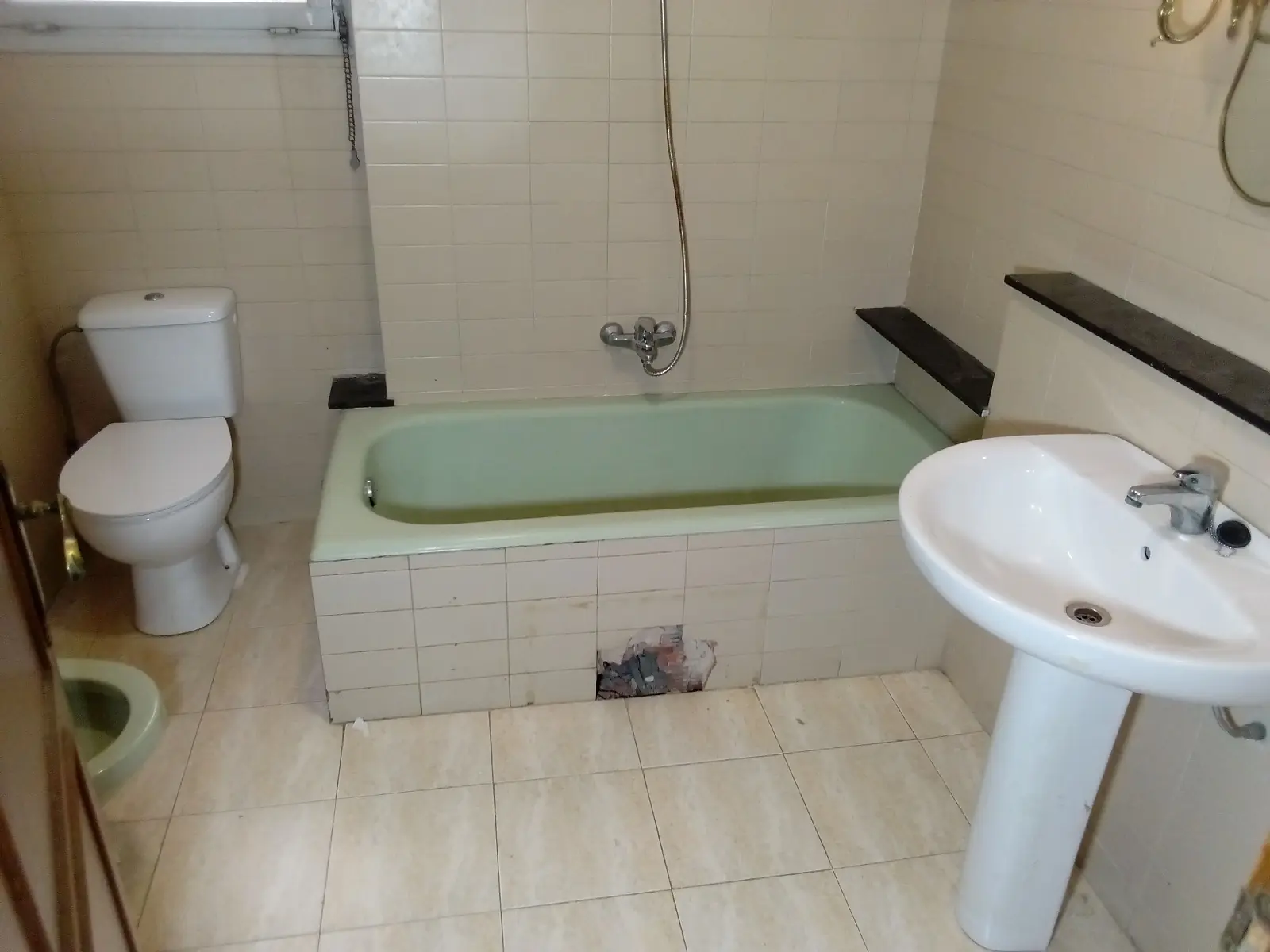 Baño antiguo con bañera verde, inodoro, lavabo de pie y un azulejo roto