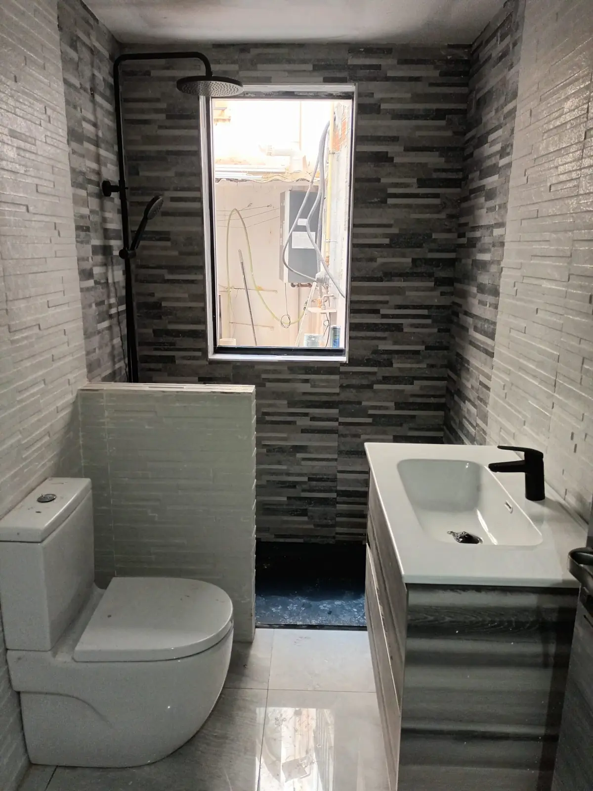 Baño reformado con inodoro, mueble de lavabo, grifería negra y ducha bajo una ventana