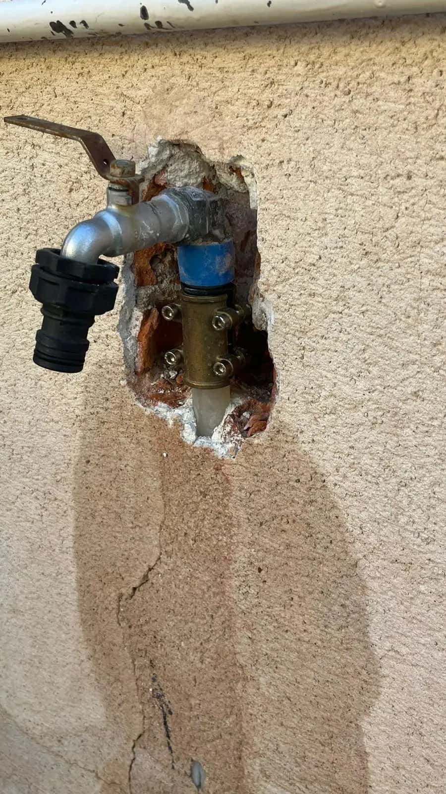 Grifo exterior con la pared picada, abrazadera en la tubería y mancha de agua debajo