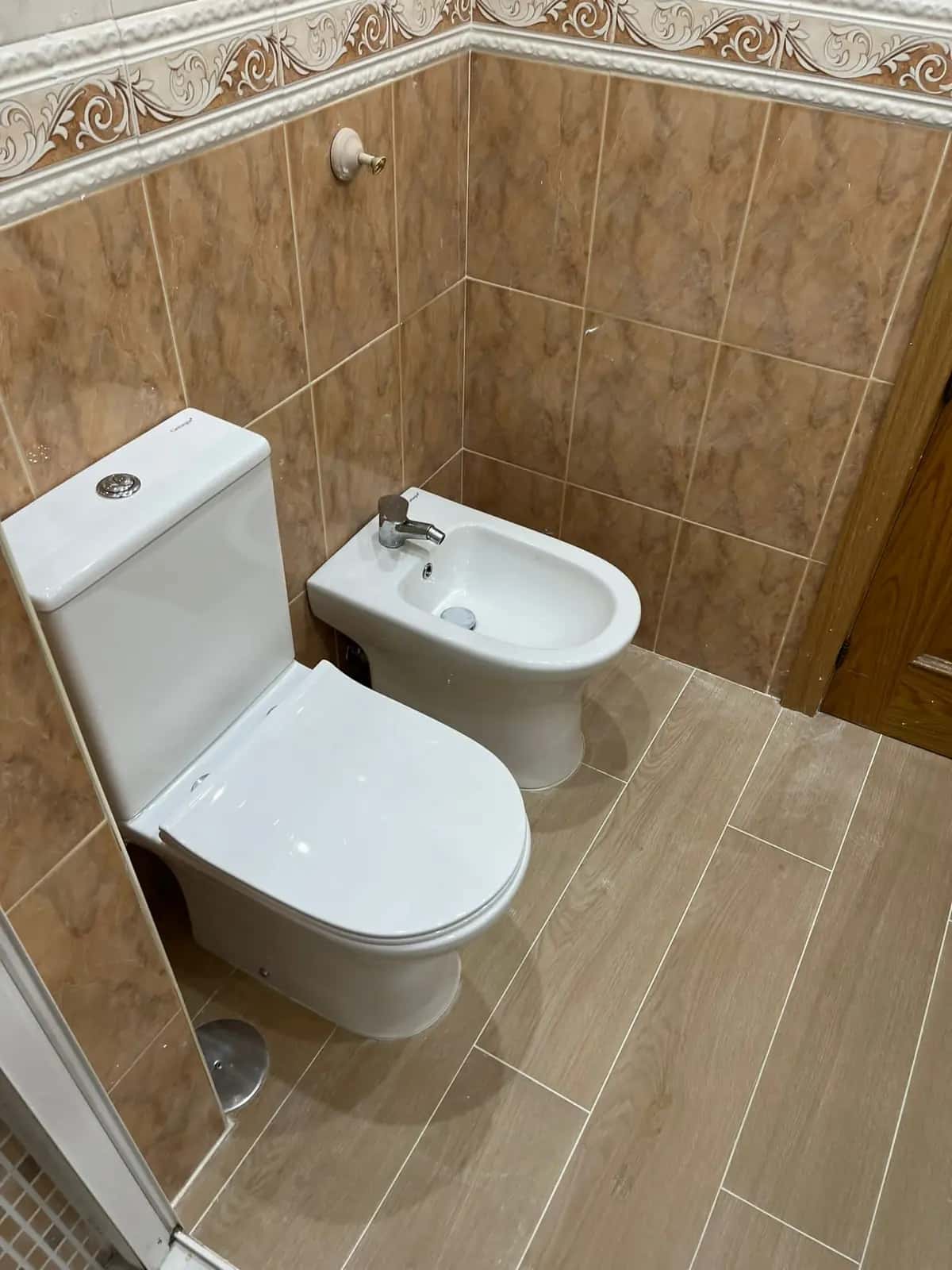 Inodoro y bidé blancos nuevos en un baño con azulejos marrones y suelo imitación madera