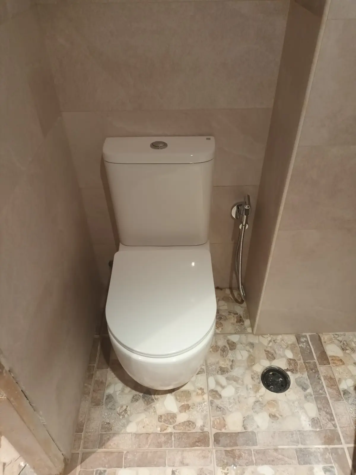 Inodoro blanco con grifo de higiene junto a un sumidero en suelo de piedra
