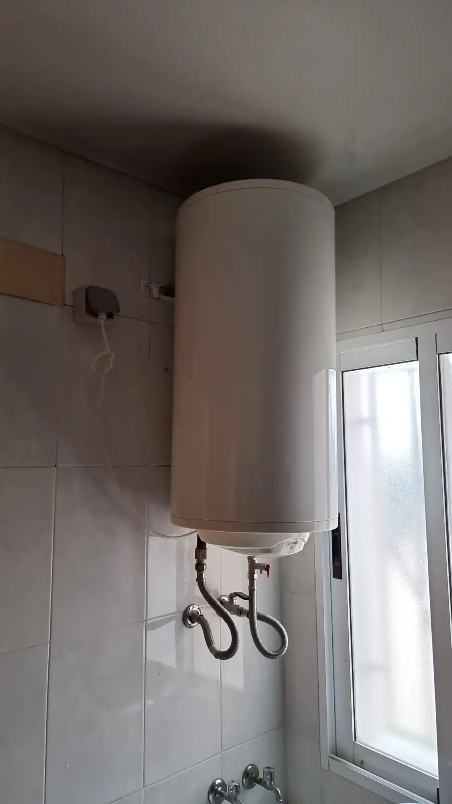 Termo eléctrico colgado junto a una ventana con latiguillos de conexión a la vista