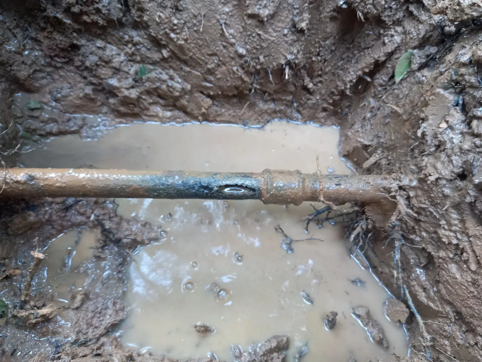 Tubería enterrada con una fuga de agua en una zanja embarrada y encharcada