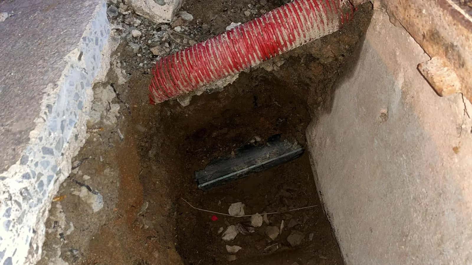 Zanja excavada junto a un muro con tubo corrugado rojo y tubería al fondo