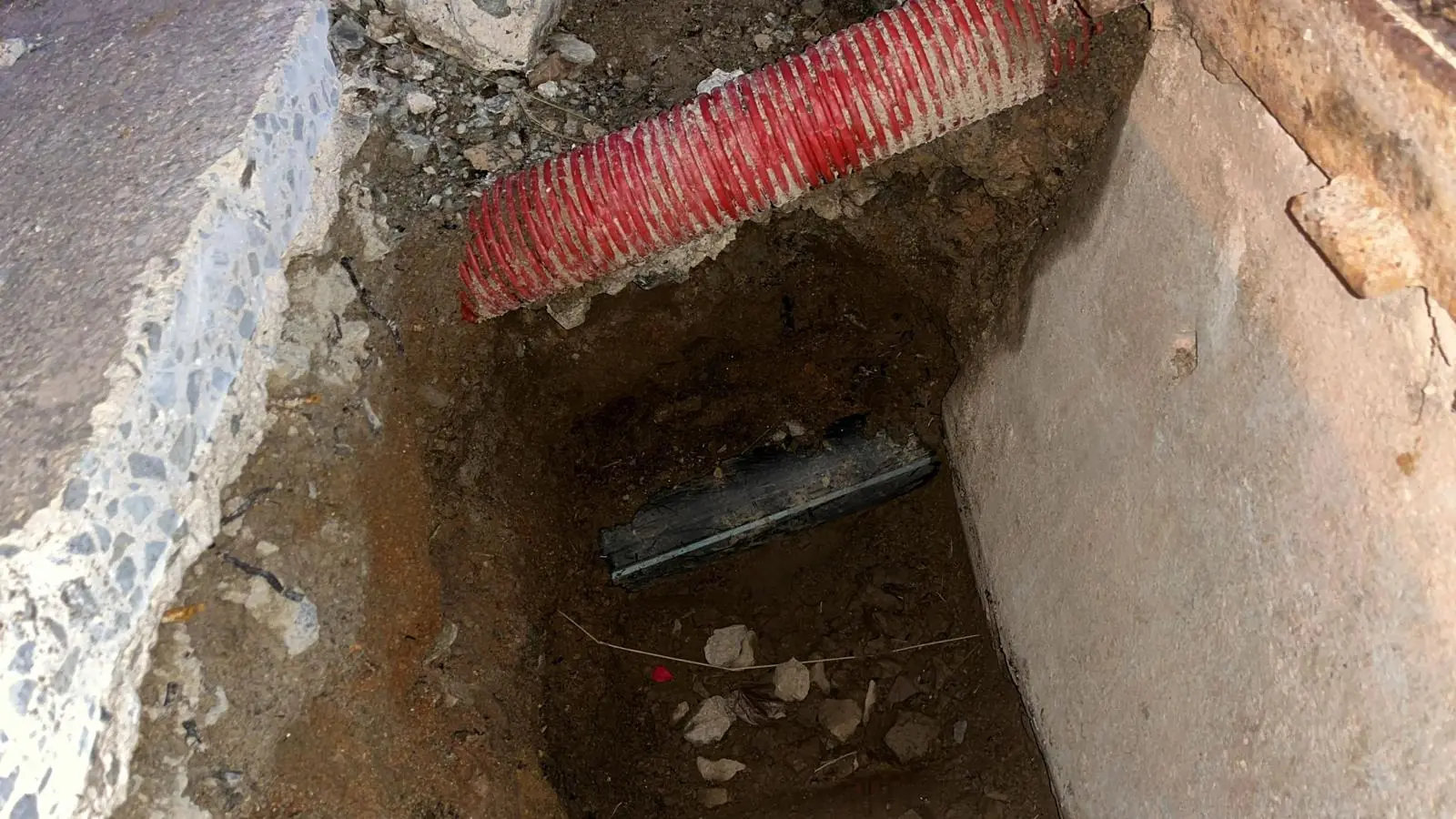 Zanja excavada junto a un muro con tubo corrugado rojo y tubería al fondo